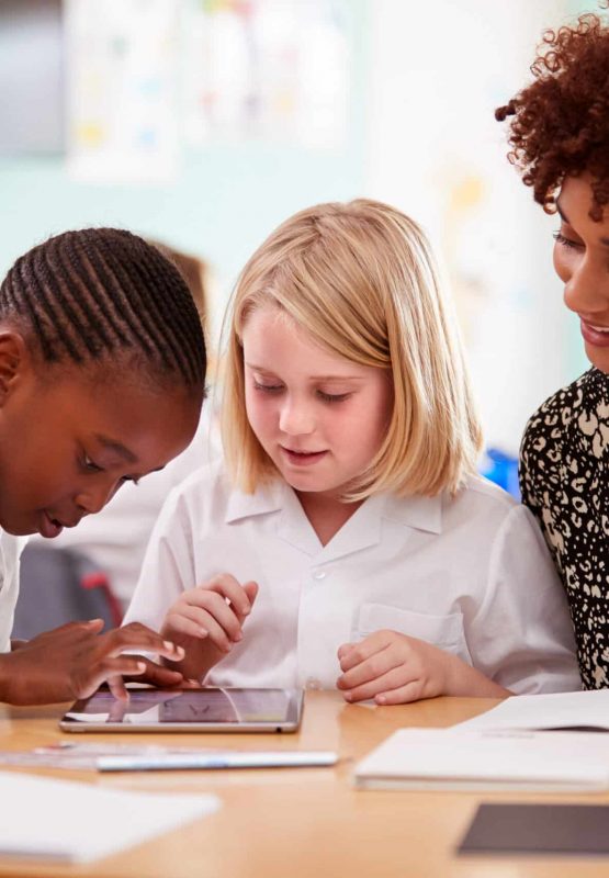 Female Teacher With Two Elementary School Pupils Wearing Uniform Using Digital Tablet At Desk Female Teacher With Two Elementary School Pupils Wearing Uniform Using Digital Tablet At Desk